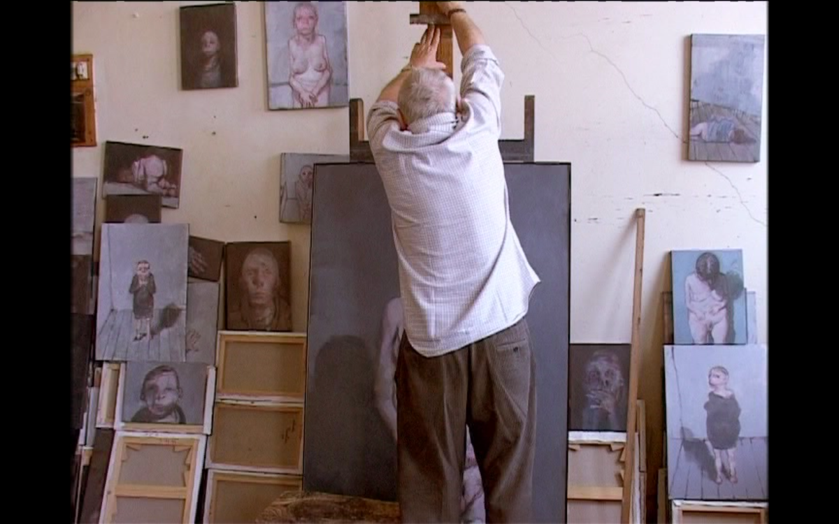 dans un atelier de peintre, un homme de dos pose un tableau sur un chevalet 