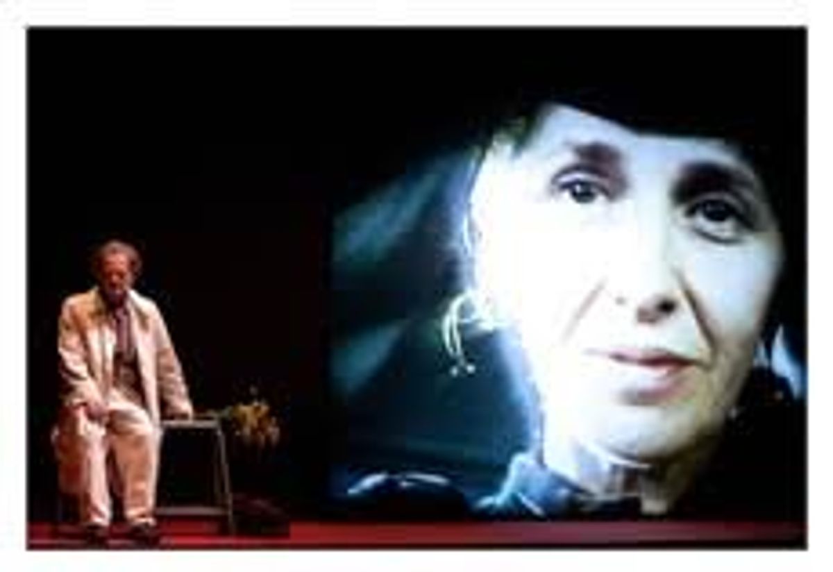 un homme sur une scène assis à l'avant plan : le visage d'une femme en vidéo sur le fond de scène